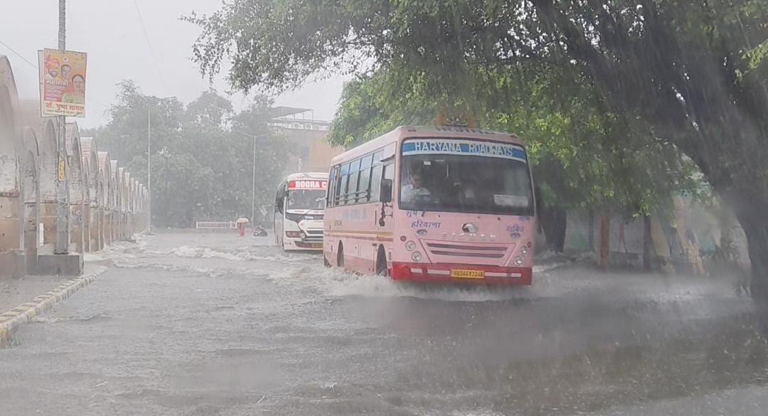 तेज बारिश के चलते शहर की सड़कों पर जमा हुए बरसाती पानी के बीच से गुजरते वाहन।